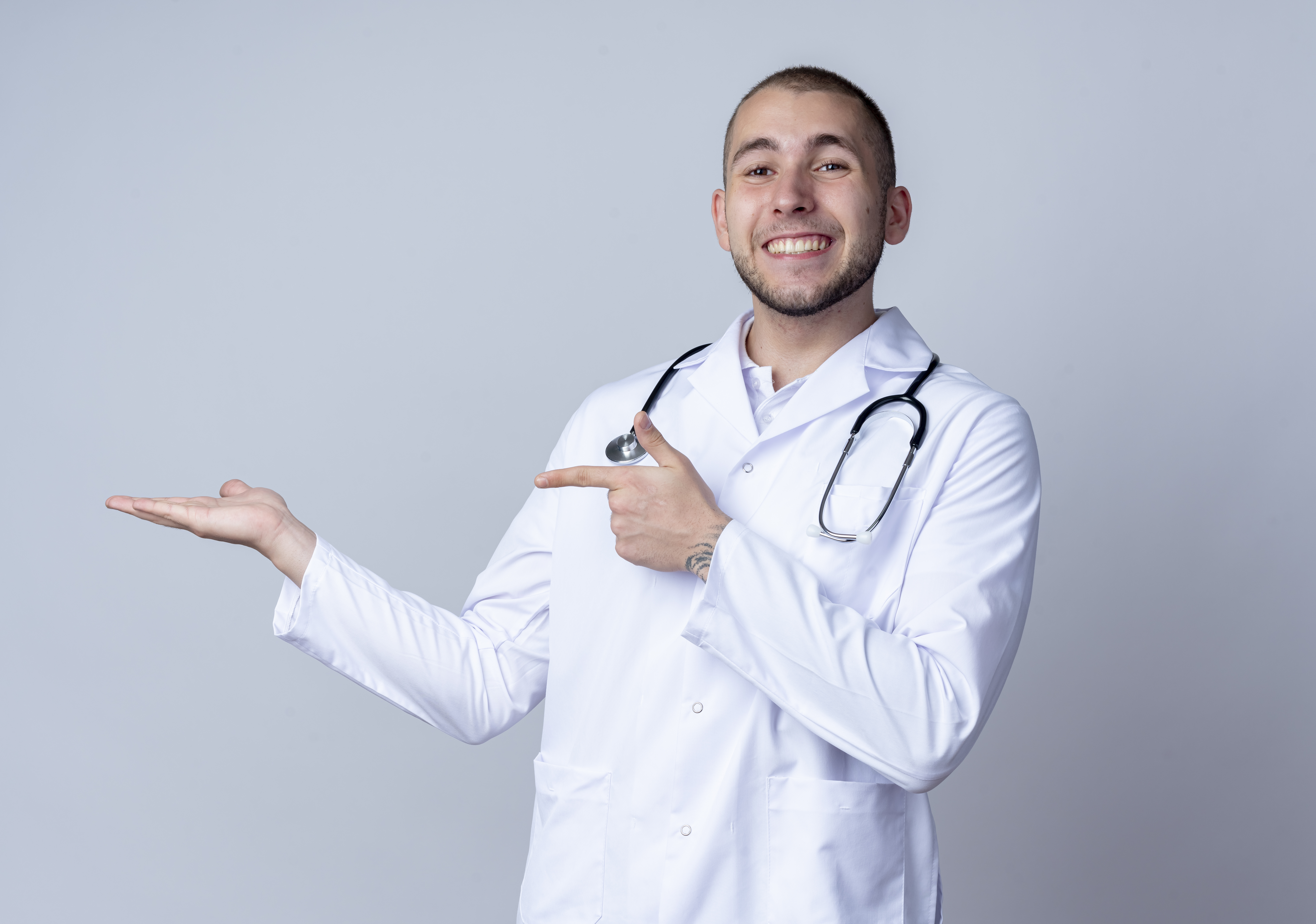 smiling young male doctor wearing medical robe and stethoscope around his neck showing empty hand and pointing at it isolated on white background smiling young male doctor wearing medical robe and stethoscope around his neck showing empty hand and pointing at it isolated on white background